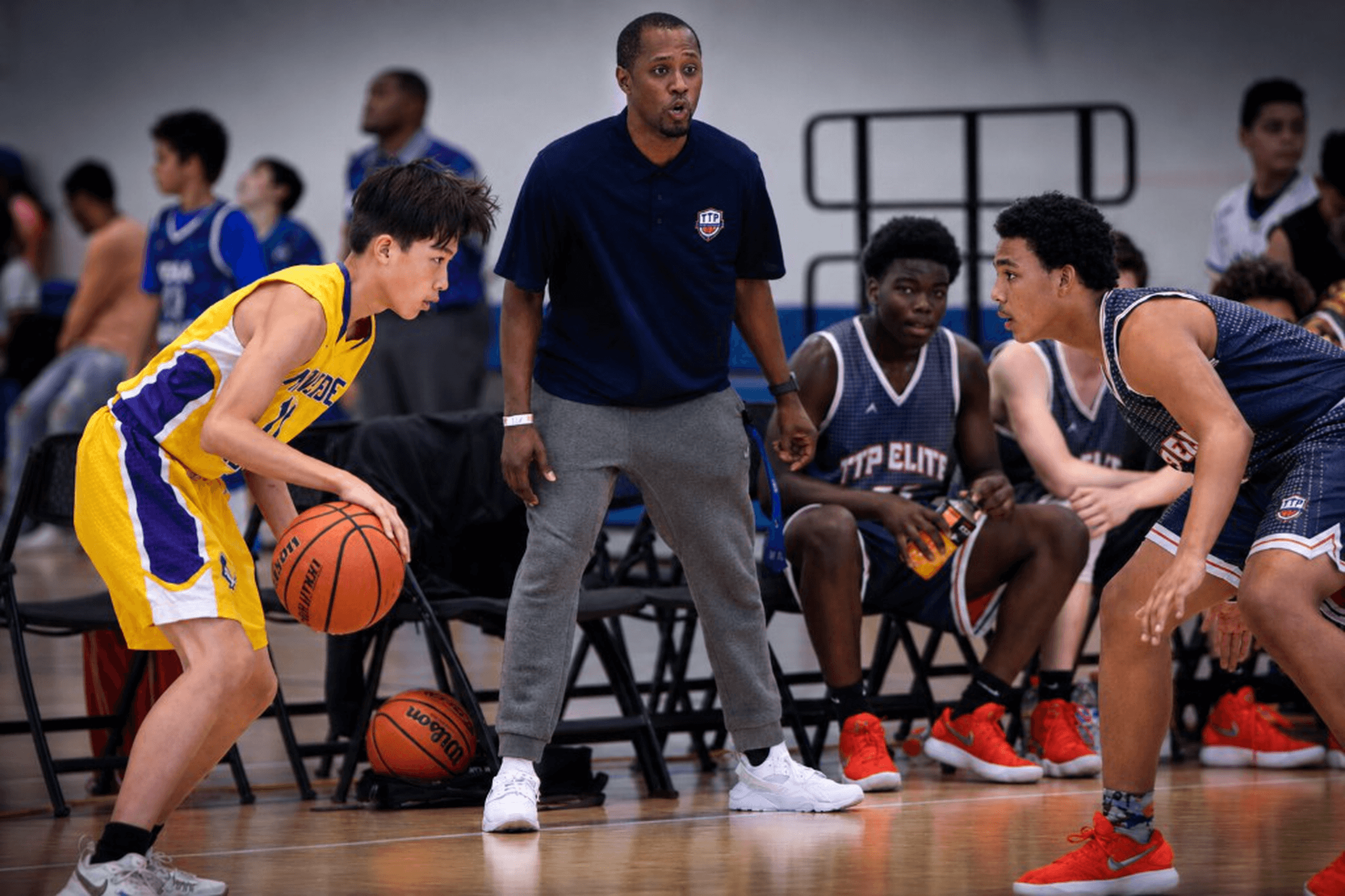Youth basketball game — yellow jersey dribbler guarded one-on-one with coach observing courtside.
